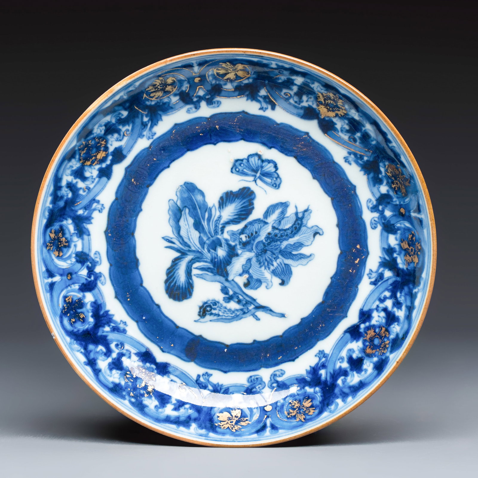 A Chinese blue and white gilt-decorated 'Botanical' plate based on designs by the German botanist: A Chinese blue and white gilt-decorated 'Botanical' plate based on designs by the German botanist Maria Sybille Merian, Qianlong, ca. 1740 Dia.: 15,5 cm Decorated in rich underglazed cobalt blue and h