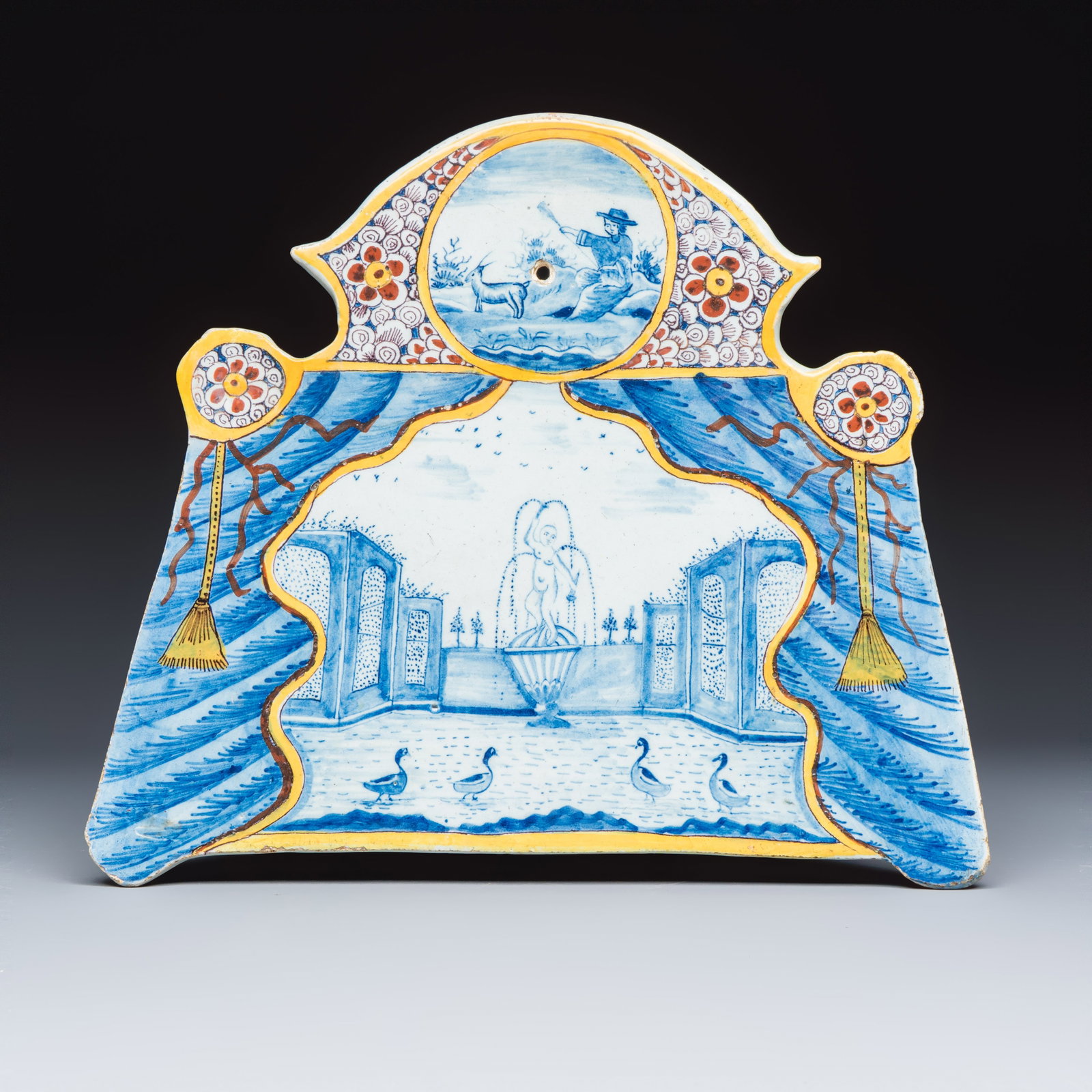 A polychrome Dutch Delft trompe-l'oeil plaque with a fountain garden in a theatrical curtain frame, (1 of 2)