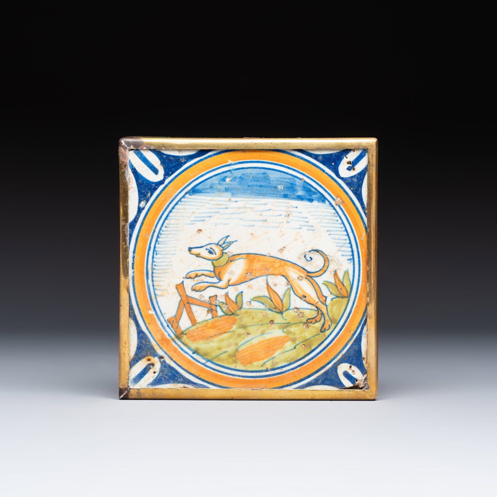 A polychrome Dutch Delft maiolica medallion tile with a leaping dog, 16th C.: A polychrome Dutch Delft maiolica medallion tile with a leaping dog, 16th C. Dim: 13,6 x 13,6 x 2,9 cm | incl. mount Mounted in a brass frame for suspension or use as a coaster.