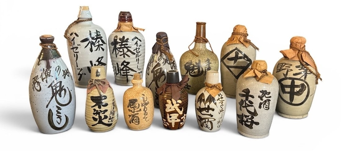 A Collection of Japanese Tokkuri Sake Bottles: 20th Century. A collection of 12 bottles. Most seem to be unused. Some with corks, some with cups, some with both. Some bottles retain original labels. Tallest measures 12.5" H