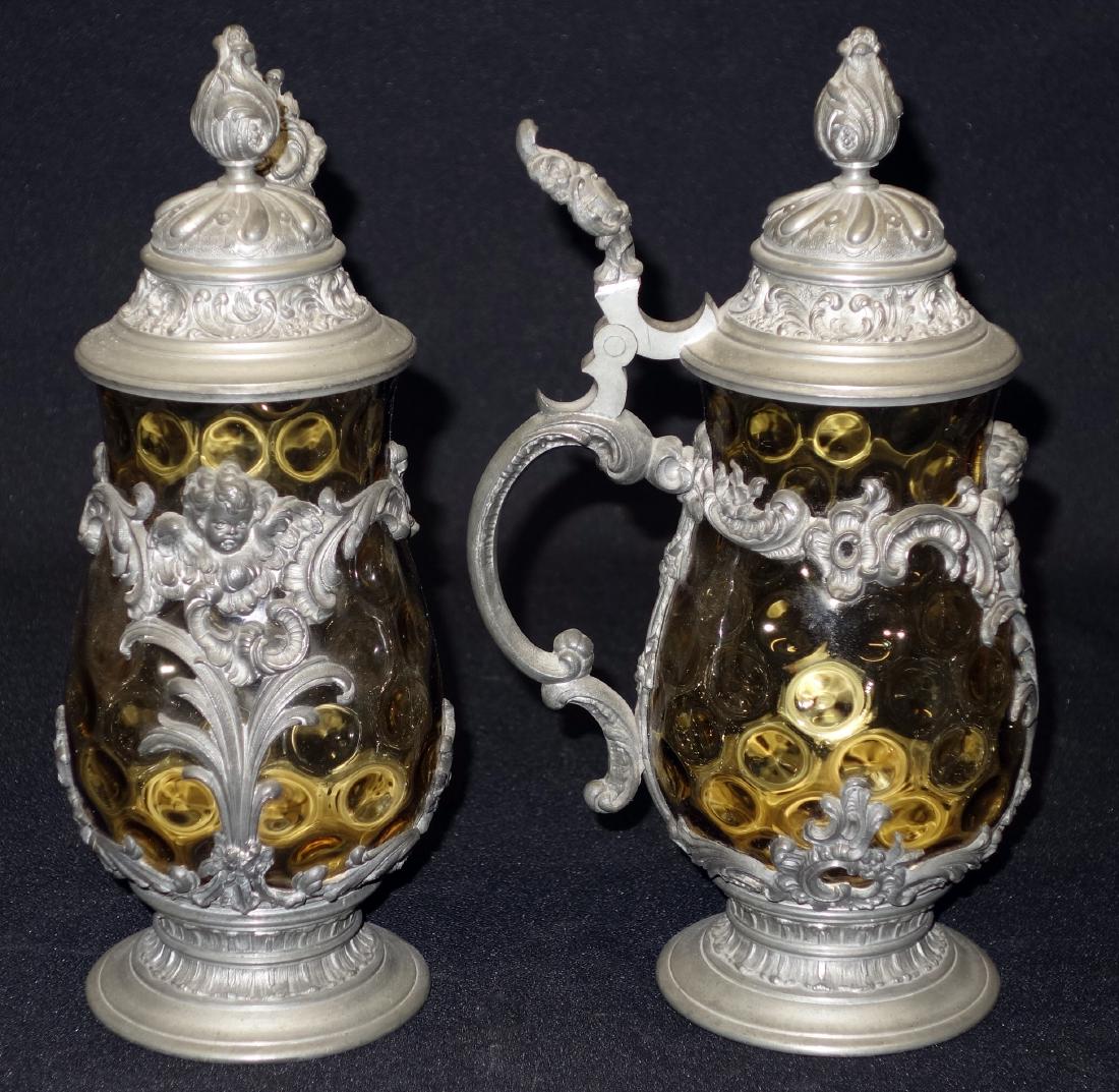 PAIR OF GLASS STEINS: Amber thumbprint glass surrounded by pewter shell with cherubs and foliate decoration; normal wear; H-9 1/2", G-VG