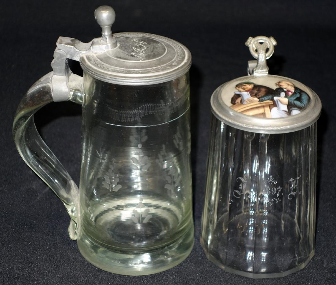 GLASS STEINS (2): Pewter lids; (1) has painted porcelain with saying; (1) .5 L; paneled; 1897; painted porcelain top with saying; (1) etched monogram lid; H- 6 1/2- 8"; V-G