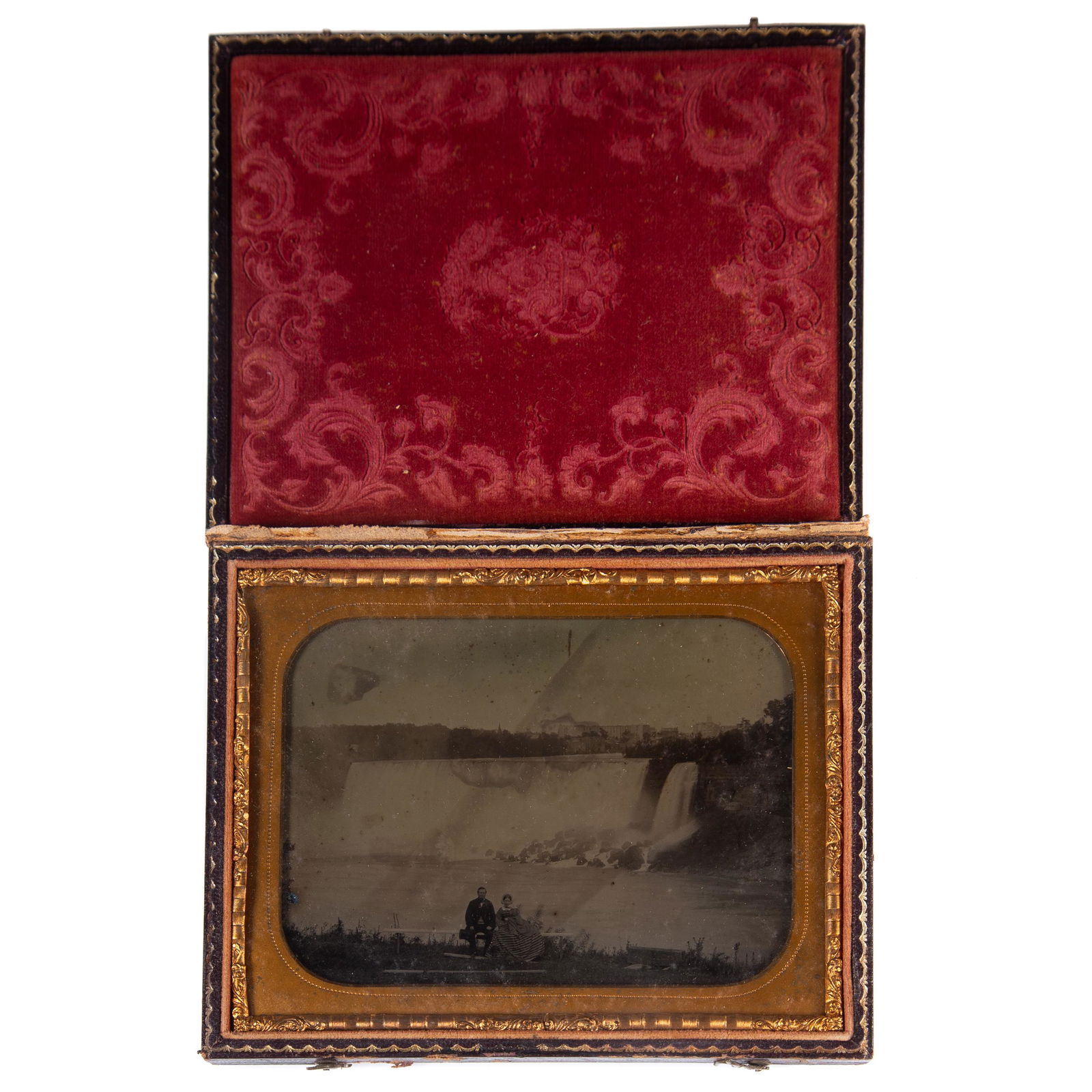 AMBROTYPE NIAGARA FALLS: Half plate; image of well dressed middle aged couple seated on bench in front of falls; man holding top hat; simple gilt mat; stamped grape and urn leather case; case hinged separated; some marks in i