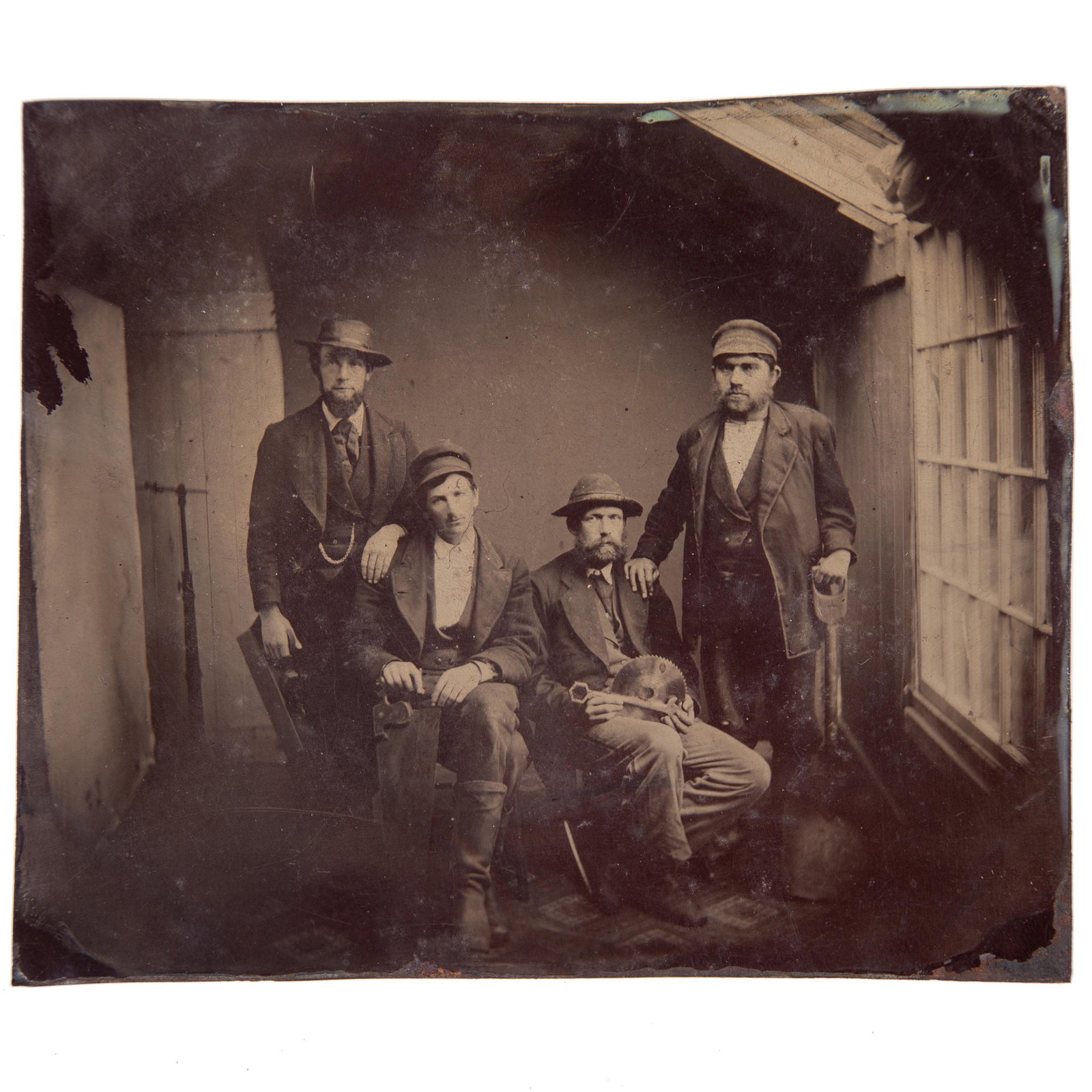 OCCUPATIONAL TINTYPE: Half plate; image of (4) carpenters, two seated, two standing; all wearing hats; holding tools including: wood plane, hand saw, wrench, circular saw blade, shovel; unusual in that the studio room has 