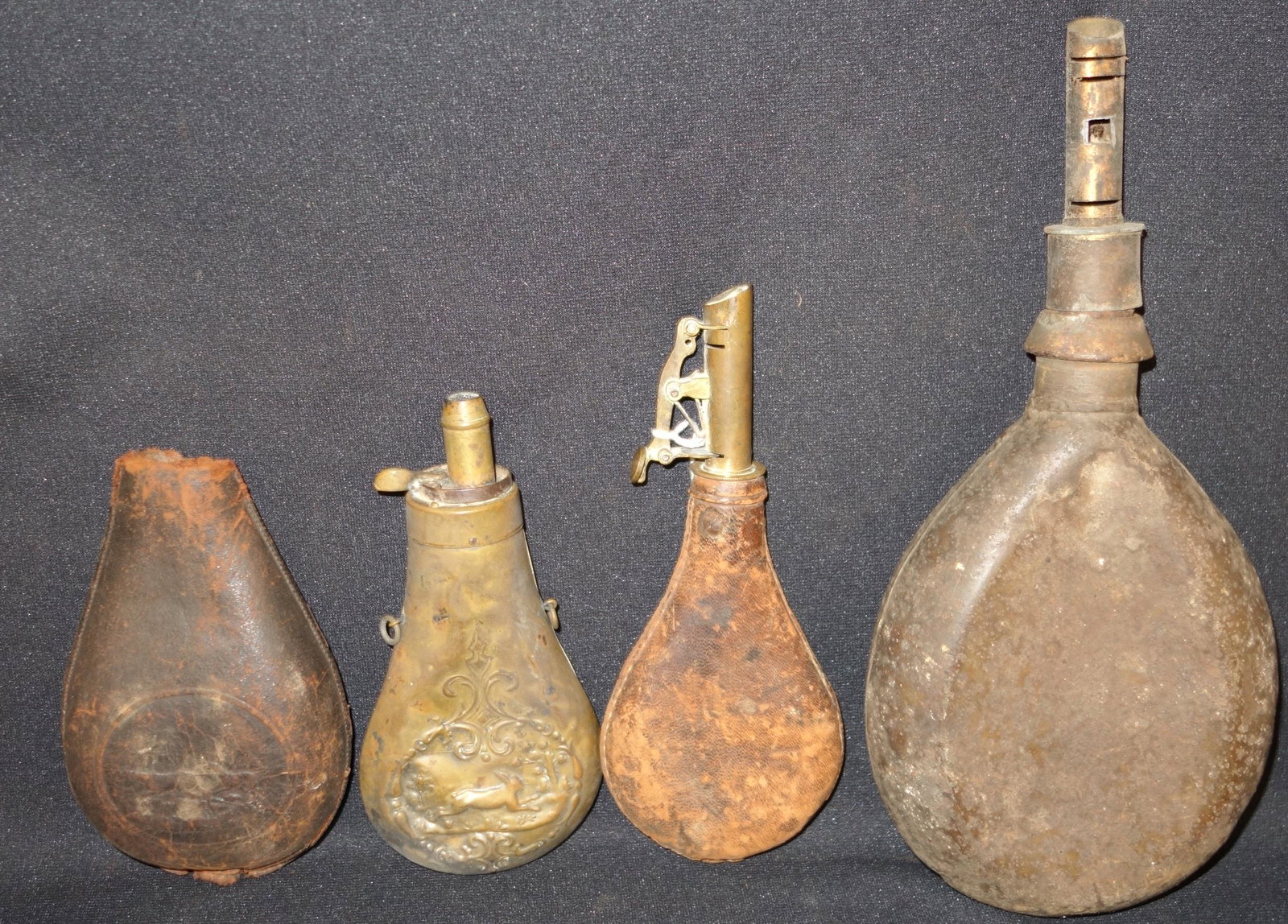 ANTIQUE POWDER FLASKS (4): Metal, brass and leather; (1) brass, dented, embossed rabbit scene on both sides; (2) leather, 1-side seam is open and has some embossing; (1) metal, possibly a canteen; heavy; heavy wear overall; lon