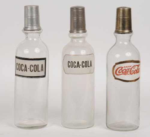 3 ASSORTED COCA-COLA SYRUP BOTTLES: All stand approximately 12" tall; the1920's bottle with the foil label is strong overall with only some dark marks on the right side of the label and a small chip concealed by the cap on the top of th