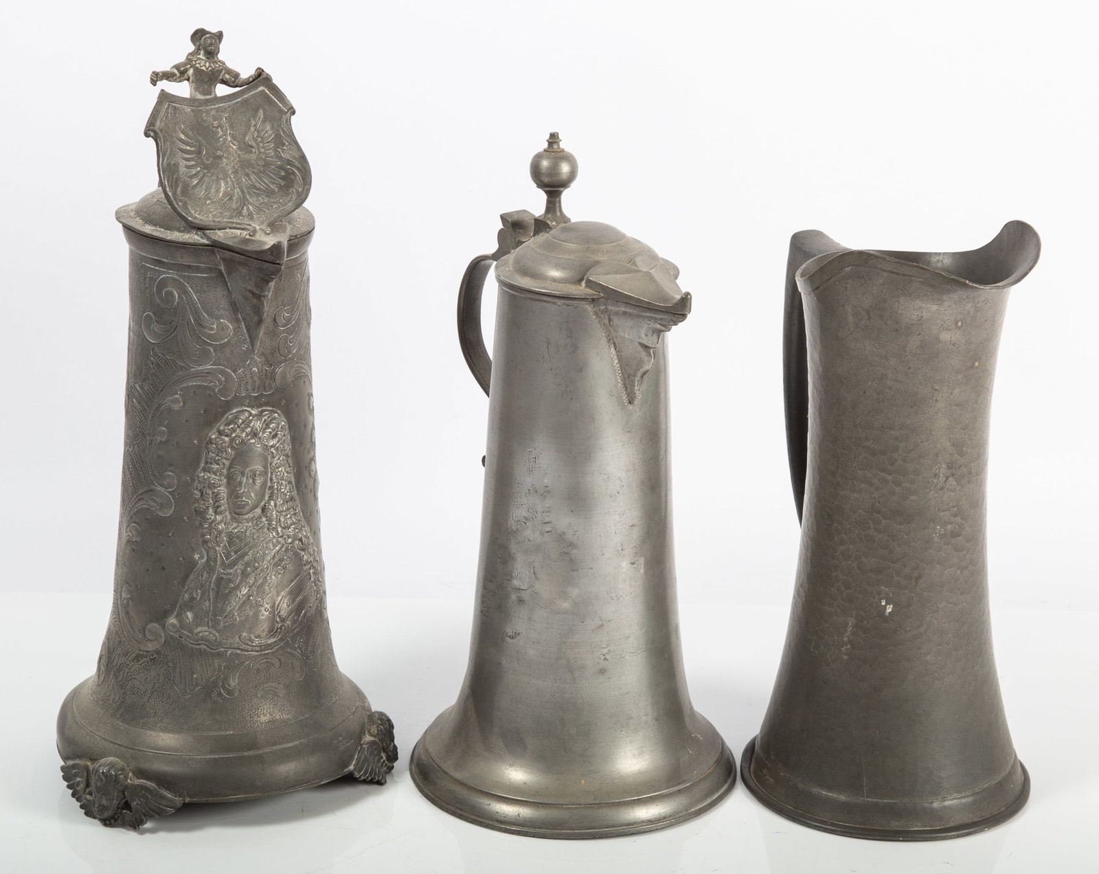 PEWTER TANKARD PITCHERS (3): (1) Highly embossed with royalty figure, shield finial on hinged lid, cherub feet; (1) plain with ball finial on hinged lid; (1) hammered pitcher, no lid, 20th Century; dings, dents and wear; H-10 1/2