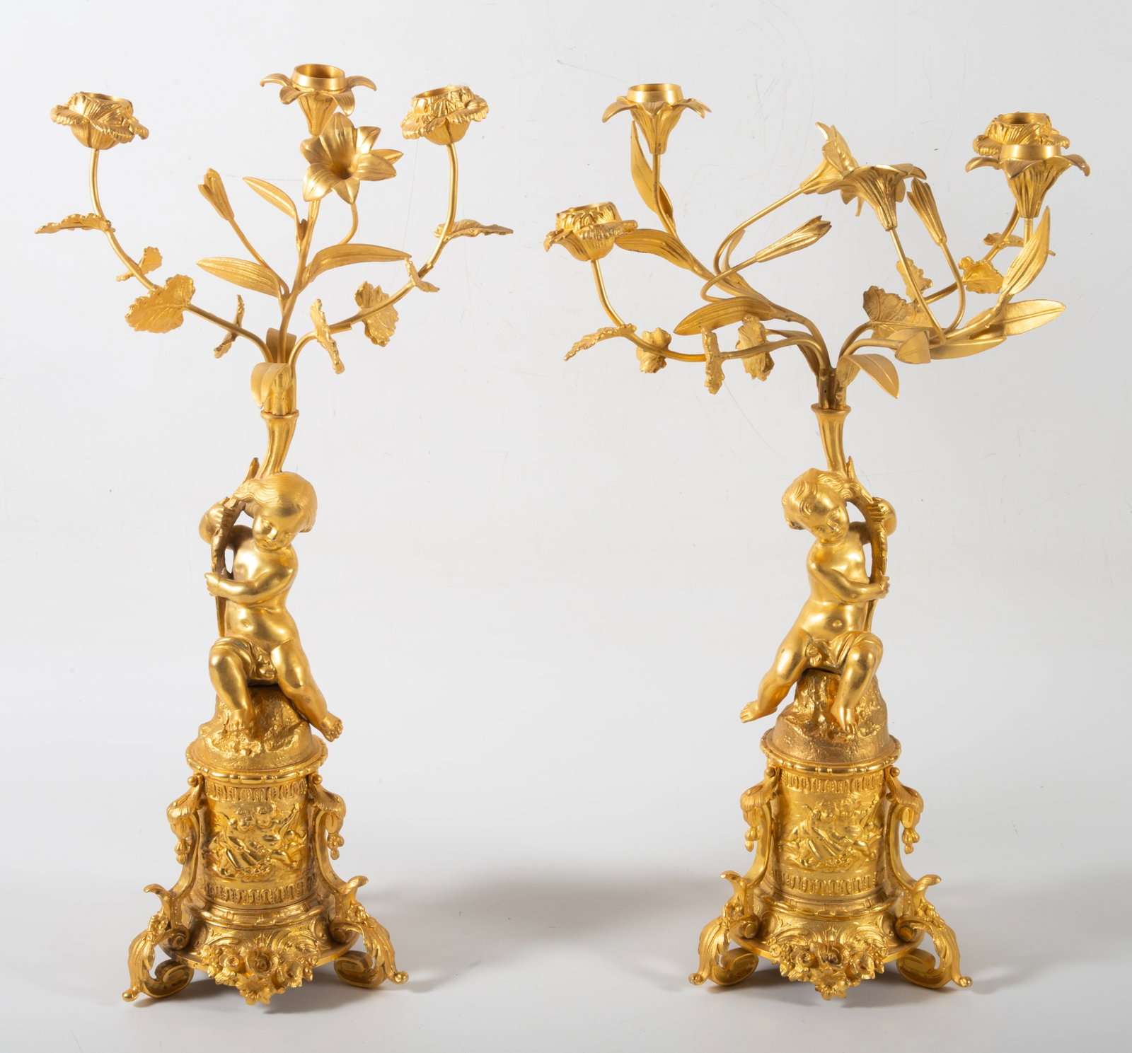 GILT BRONZE CANDELABRA: Pair of gilt bronze column form candelabras- figural cherubs holding four cup floral candle arms, ornate base with three legs, flowers and embossed cherub scenes; normal wear, one candle cup and arm