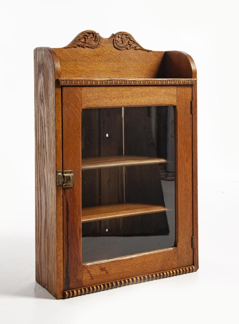 Victorian Oak Hanging Wall Cabinet: DESCRIPTION: Victorian oak hanging wall cabinet. Single pane glass door and two shelves. Broken arch top with carved foliate detail. Brass closure. MEASUREMENTS: 13-3/4" wide x 7" deep x 32-1/2" high.