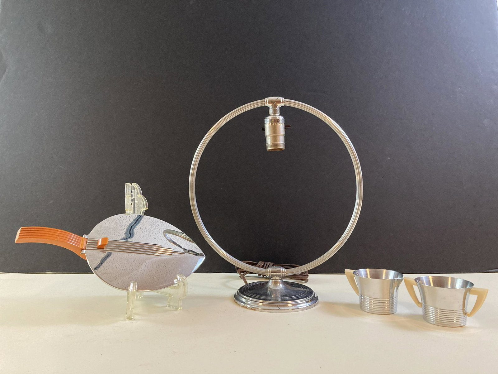 3 Chase Chrome Articles: Chrome Lamp Base, Silent Butler with Bakelite, Cream and Sugar dishes. Lamp Base 12 “ wide x 14 “ high. Silent Butler 6“ wide x 11 1/2“ high. Cream and Sugar Dishes 3“ diameter x 2“ high. 