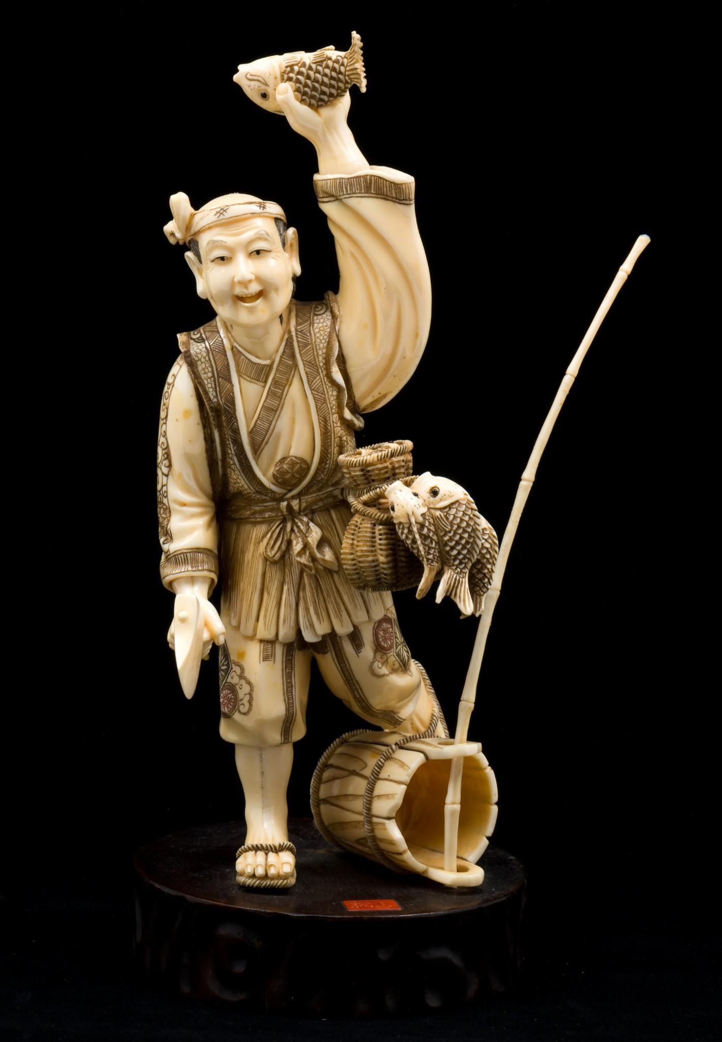 Japanese Okimono Of A Fisherman: Carved okimono of a fisherman holding a fish and an axe. One leg lifted resting on an over turned bucket. Elaborately decorated with black and brown accents. Fixed to wood base. Chop mark to front cen