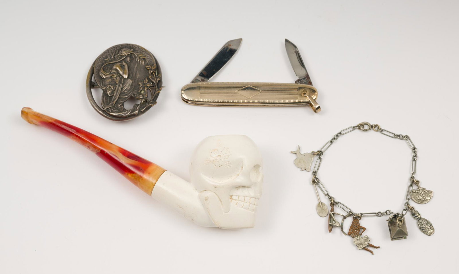 Pipe, Pen Knife, Bracelet & Brooch: Includes faux Meerschaum pipe of skull, The Ideal company penknife, base metal brooch of seated woman with flowers surrounding, and Hawaiian charm bracelet. Pipe 1-3/4" high x 5-1/8" long. Knife 