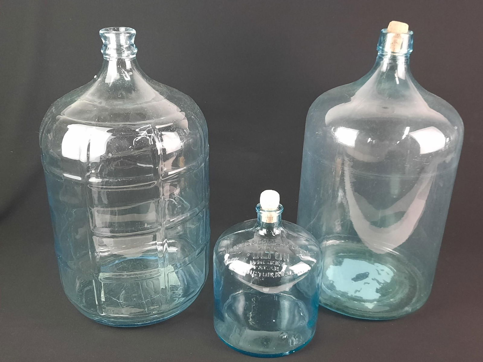 3 Vintage Glass Jugs incl Fulton Whiskey: One embossed "CRISA BOTTLE" under base. 7" to 10" diameter x 10" to 19" high.