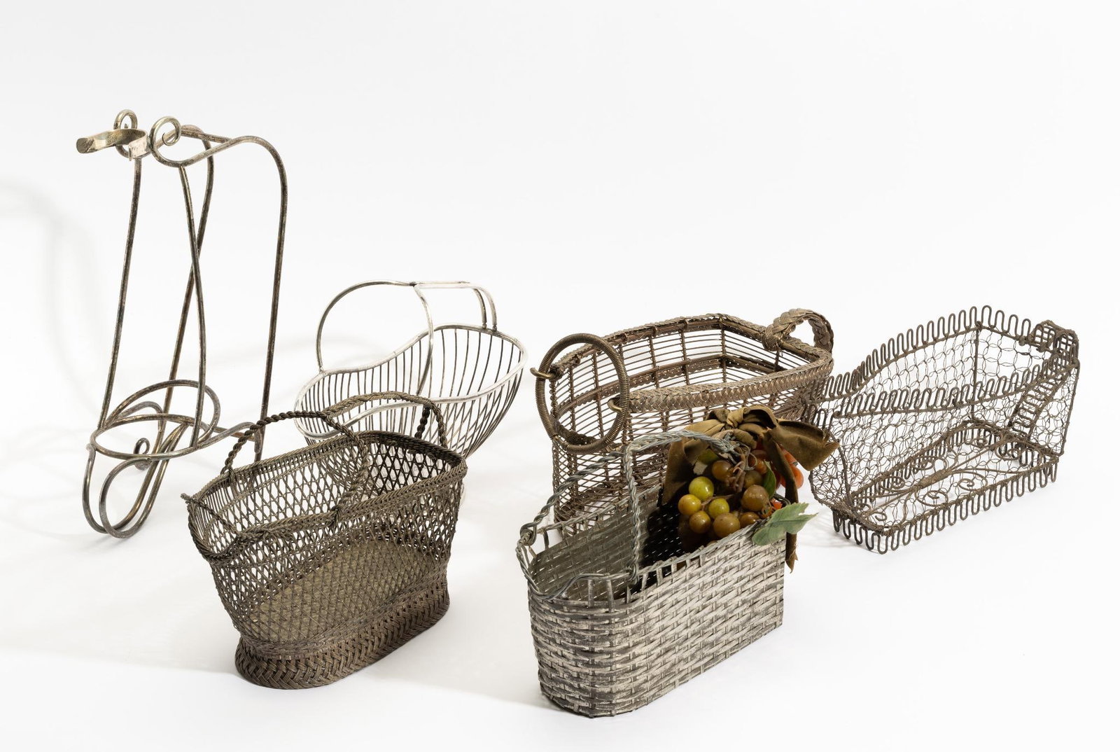 6 Antique Wine Bottle Holders: Woven wire, woven silverplated wire. The oldest an early 1800s twisted wire piece. The more recent pieces include a 20th century holder with faux fruit and the basket woven from ribbon metal. One a we