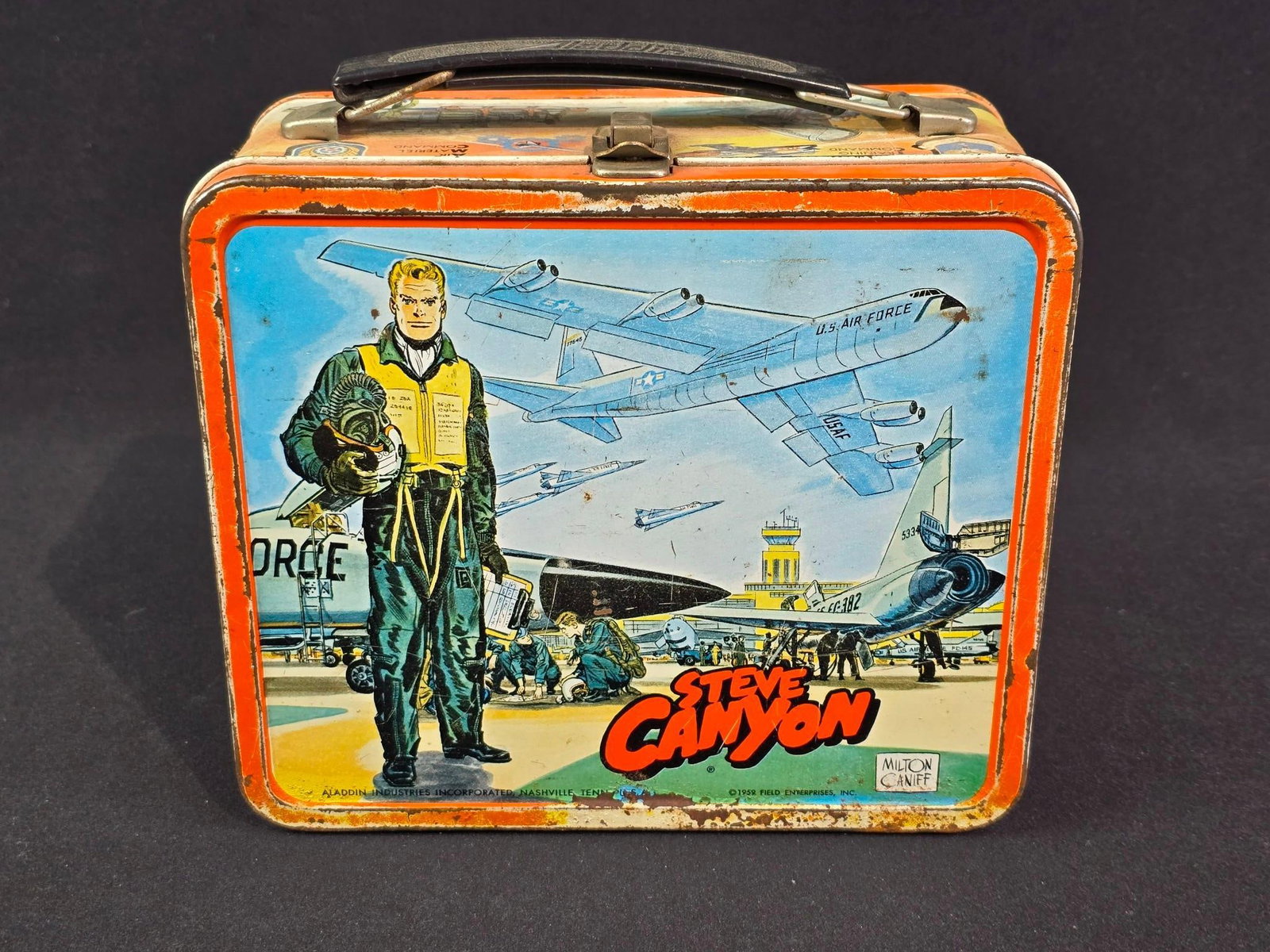 1959 Steve Canyon Lunchbox: Includes thermos. Milton Caniff artwork. 8-3/4" wide x 4" deep x 7-1/2" high. Various states of paint losses, scratches, and rust.