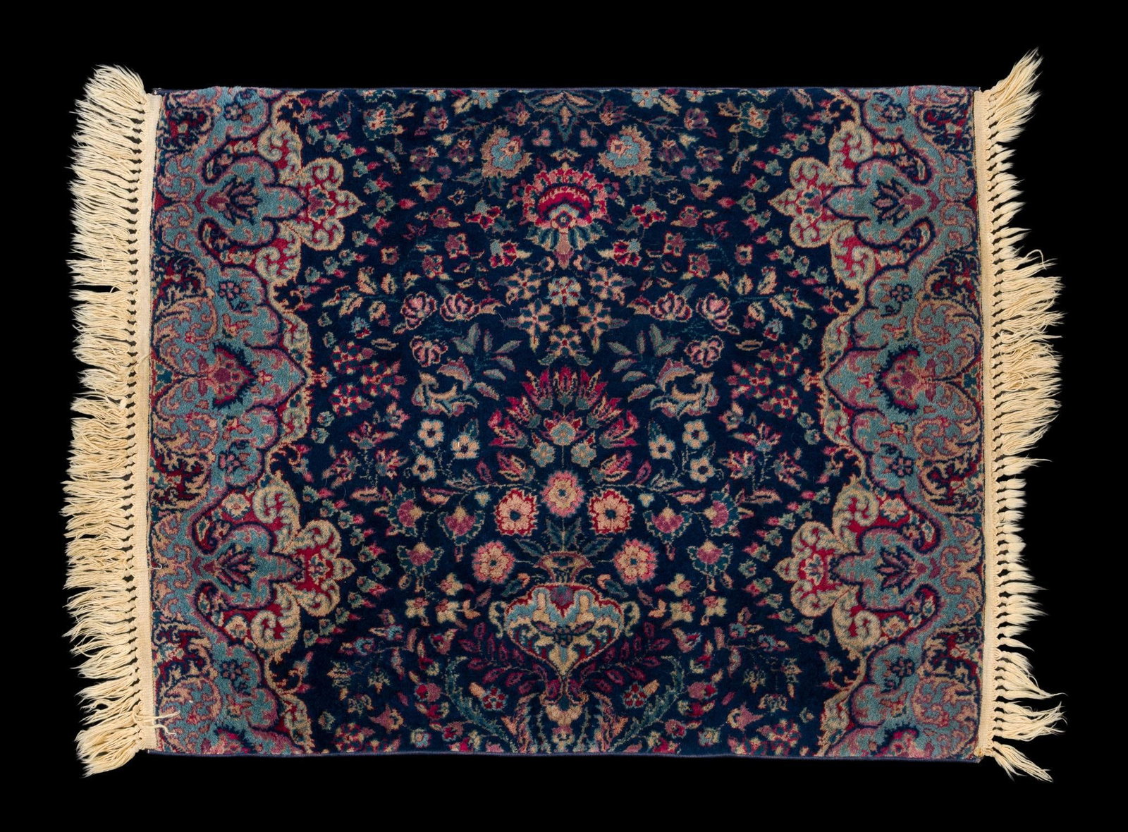 Persian-Style Kerman Design Rug: Machine-woven rug featuring a navy field with an allover floral and foliate pattern, centered on a vase-form floral arrangement, within a light blue scrolling foliate border. Finished with fringe ends