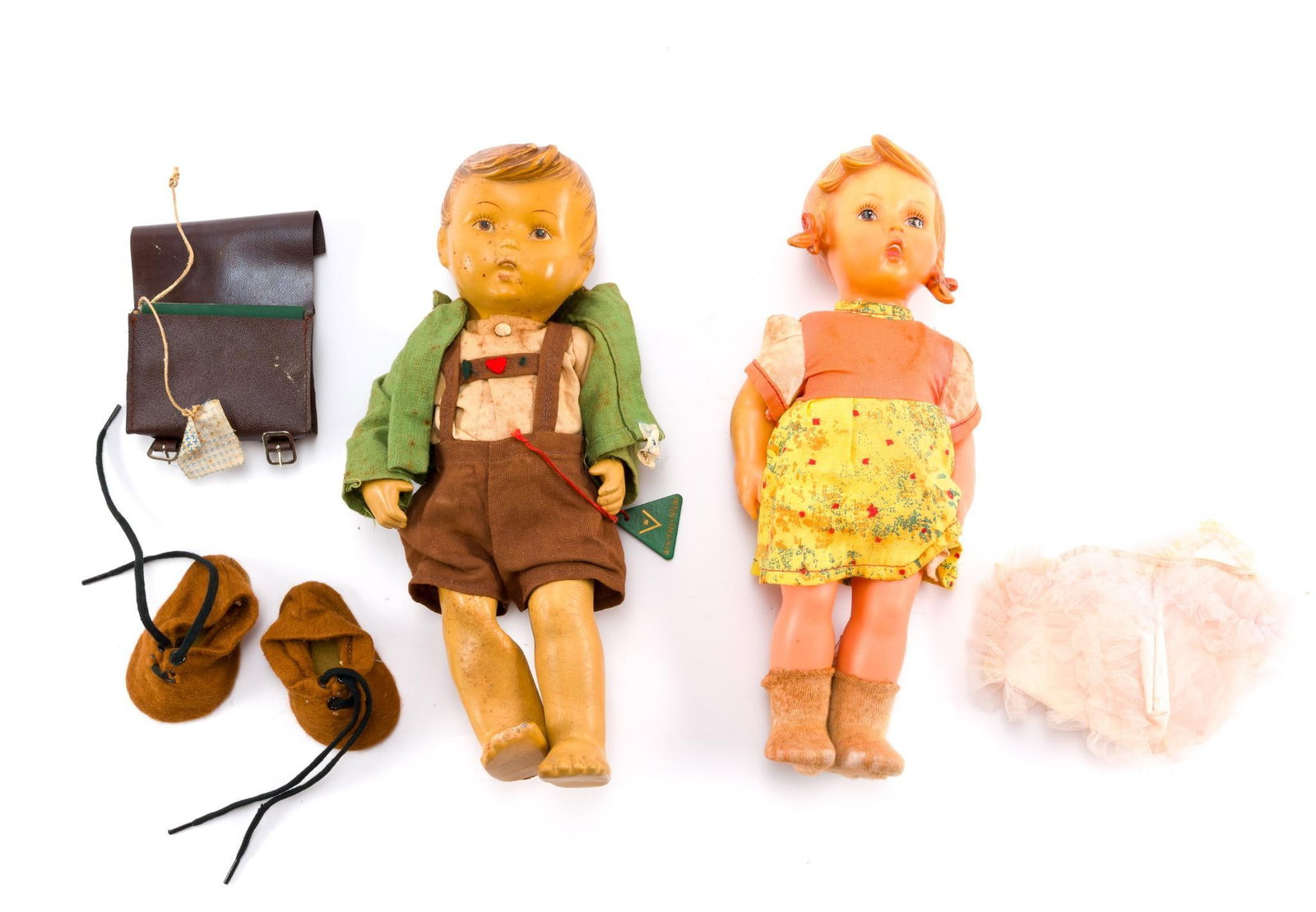 2 Vintage West German Made Dolls: Includes 2 vintage M. J. Hummel Dolls, a boy and a girl, a pair of doll shoes, a small briefcase, and a doll bonnet. In condition consistent with age and use. The boy's right leg is smashed, and the p