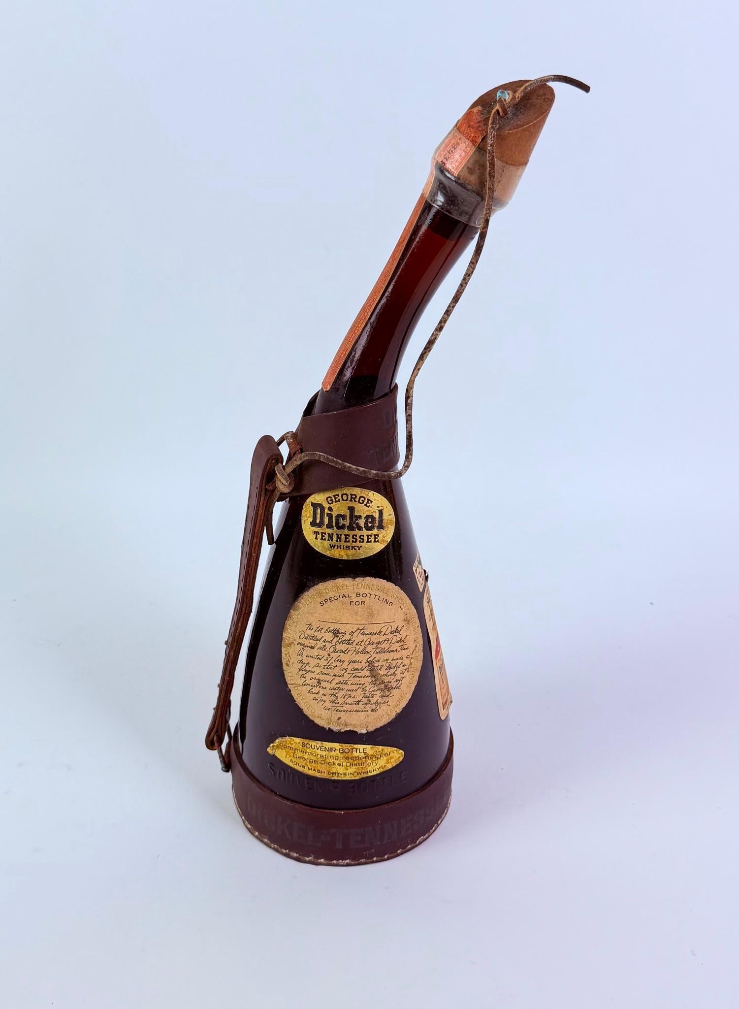 George Dickel Tennessee Whiskey: In souvenir bottle commemorating the reopening of George Dickel Distillery. With leather carrying strap. Estate alcohol. 5" wide x 14-3/4" high. IMPORTANT NOTE: This lot cannot be shipped. Must b