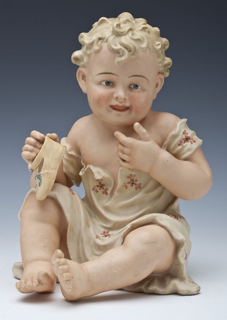 German Bisque Piano Baby: DESCRIPTION: A German bisque piano baby, probably Heubach, seated wearing a flowered garment and holding a shoe decorated with enameled dots. Unmarked. 19th century. MEASUREMENTS: 10-3/4" high. CONDIT