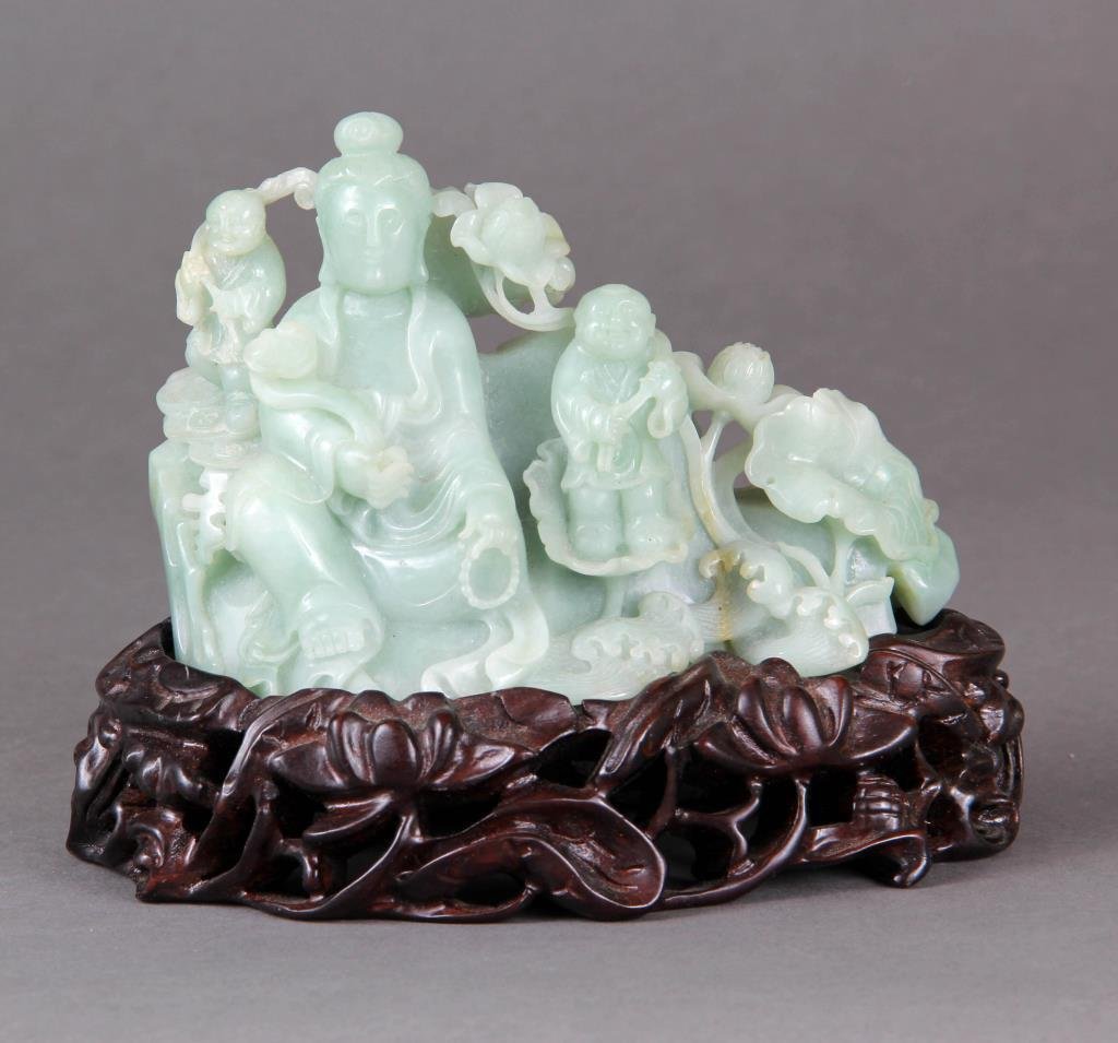 A Finely Carved Chinese Jade Sculpture: Depicting Guanyin holding a ruyi scepter surrounded by children, raised on carved hardwood stand, stone finely executed and well polished, jade measures 3.25"H x 5"W.