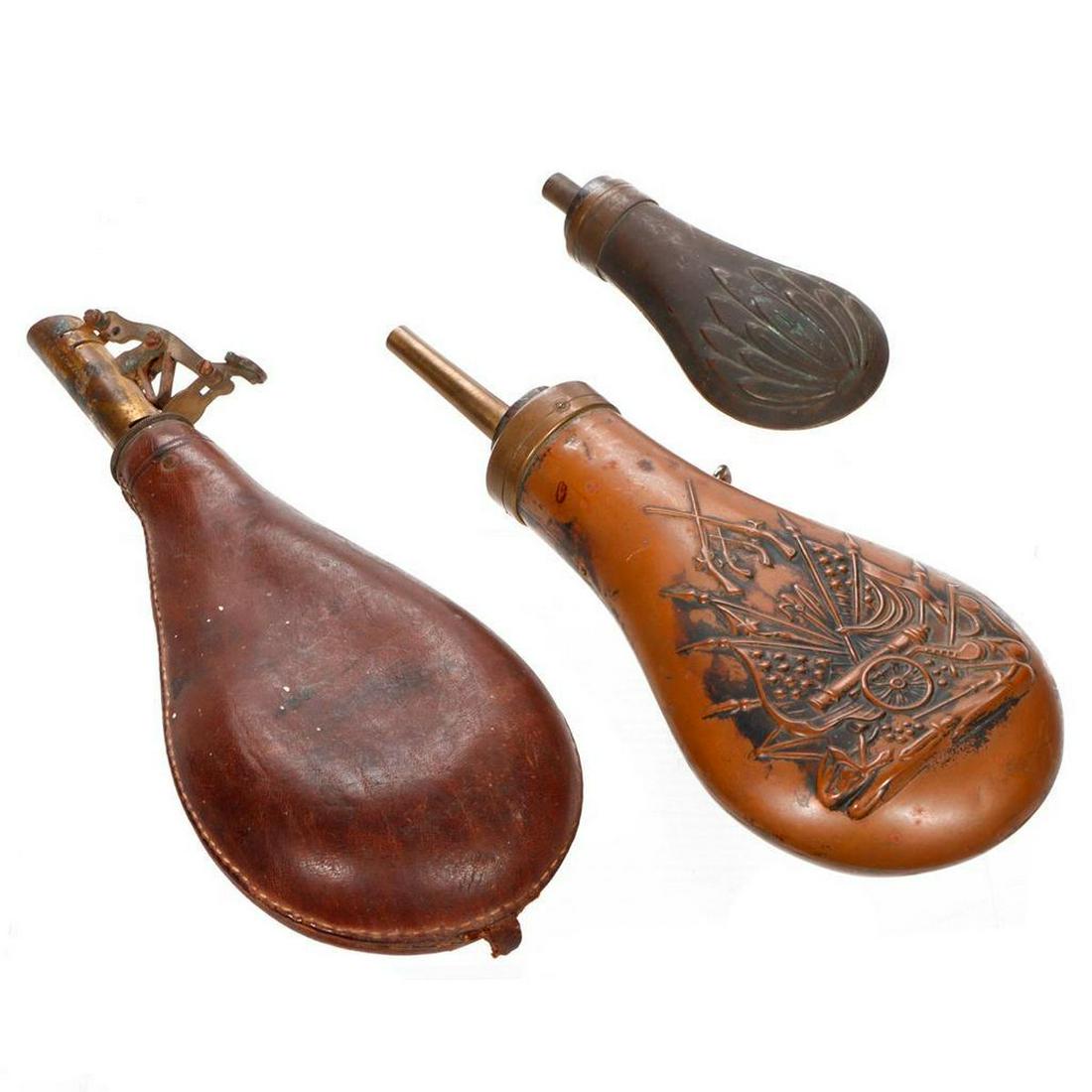 Three Vintage Powder Flasks.: Includes one small brass embossed leaf pattern (4 3/4in x 2in); one brass with commemorative flag/canon embossed decoration (8in x 3 1/2in); and one leather with brass nozzle (9in x 3 1/2in).