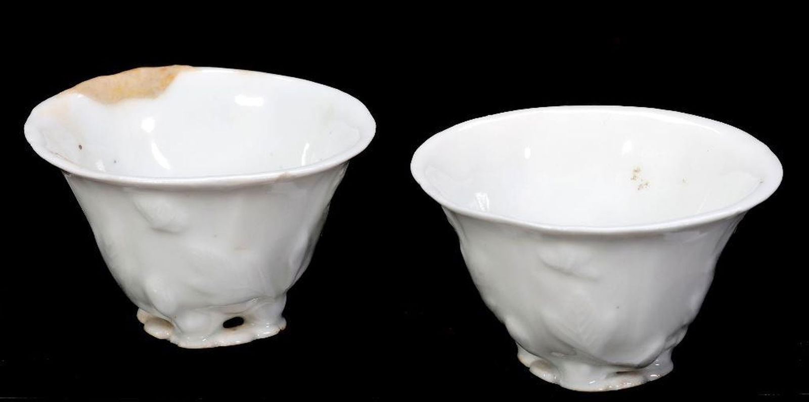 Pair Chinese Dehua Cups, 18th Century: Each small blanc de chine oval-shaped cup with molded decoration of flowers rising from foliate bases. Each approx. 2 3/8" x 2 1/8".Condition: one good; one with old repair to rim, and a tiny rim