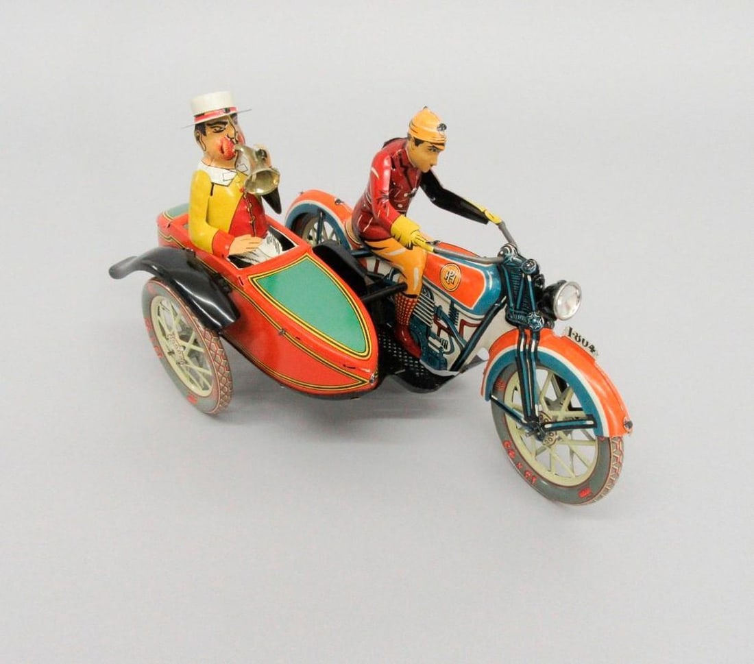 Paya S.C.V.L. Motorcycle with Sidecar: A tinplate motorcycle with side car; driver in red coat; passenger in yellow jacket with trumpet; maker's mark on exterior sides and wheels. Having the original retail box. Dimensions: 7" x 5" x 12" (