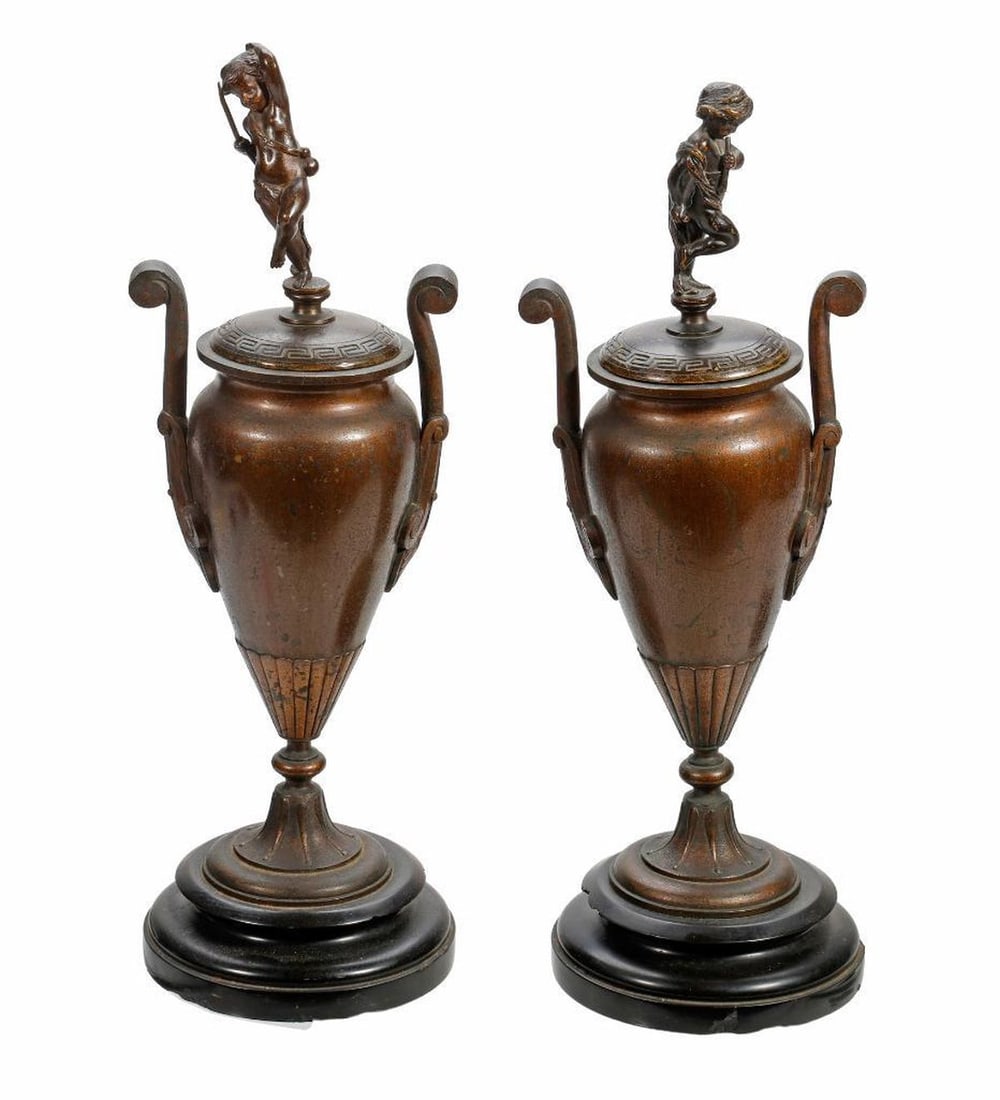 Small Pair Renaissance Revival Bronze Covered Urns: Each twin handled urn rising on a black state base, with a lid having a figural finial of nude child playing a flute, overall 11.5"h [urn slightly misshapen, chipping to the slate base]