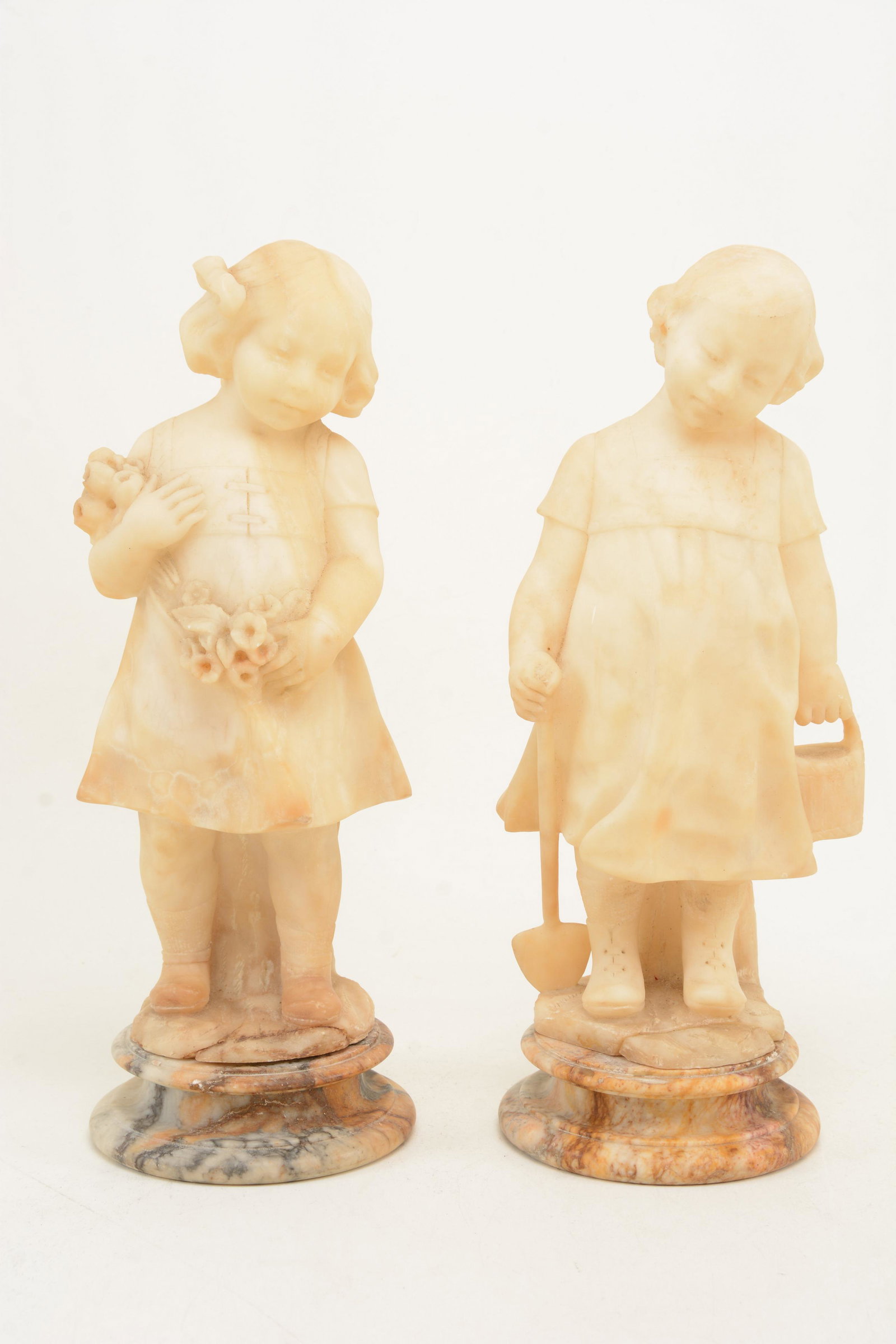 2 Alabaster sculptures of children on stands. One with: 2 Alabaster sculptures of children on stands. One with spade and bucket, the other with flowers. Some chips and losses to edges, primarily at base. Each approx.. 11in height without stands.