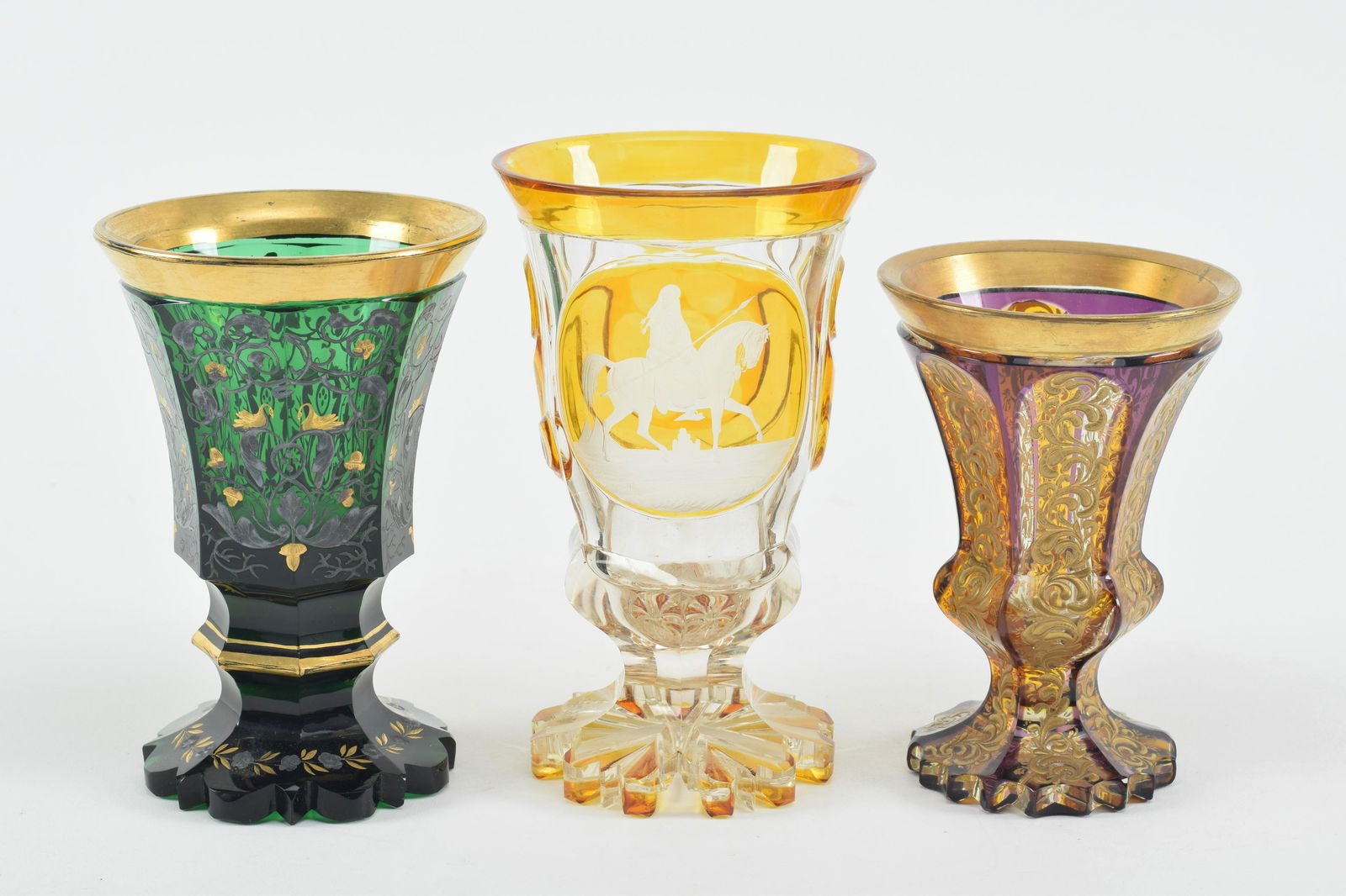 3 Bohemian glass goblets, to include a green and gilt with swans, purple and gilt with gilt scrolls: 3 Bohemian glass goblets, to include a green and gilt with swans, purple and gilt with gilt scrolls and amber cut to clear with warrior on horseback. Largest 6.25 in high. Each with small chips at foo
