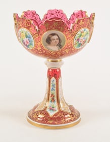 A Bohemian ruby glass and gilt tazza with floral and figural panels and a scalloped edge. Height