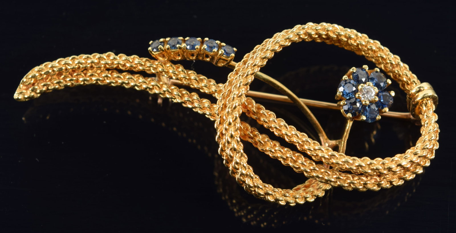 18K gold braided ribbon form pin with floral sapphire and diamond accent. Marked M 18K on reverse. 2: 18K gold braided ribbon form pin with floral sapphire and diamond accent. Marked M 18K on reverse. 2 3/8in length. 13.3 grams.