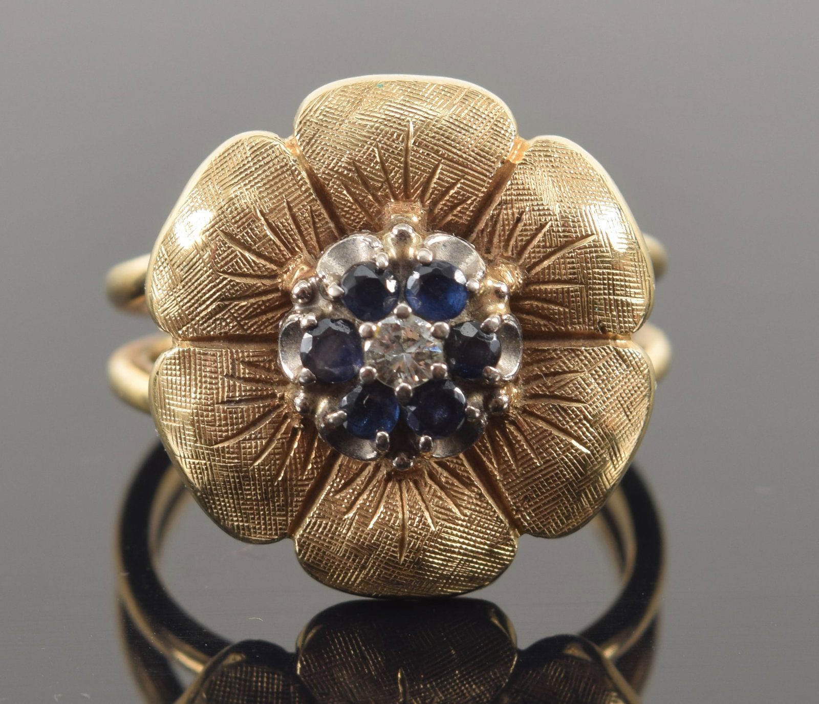 14K gold sapphire and diamond floral form ring. Central diamond (2.2mm diameter) surrounded by 6: 14K gold sapphire and diamond floral form ring. Central diamond (2.2mm diameter) surrounded by 6 round cut sapphires. Marked 14K Jabel. Ring size 5.75. 6.3 grams.