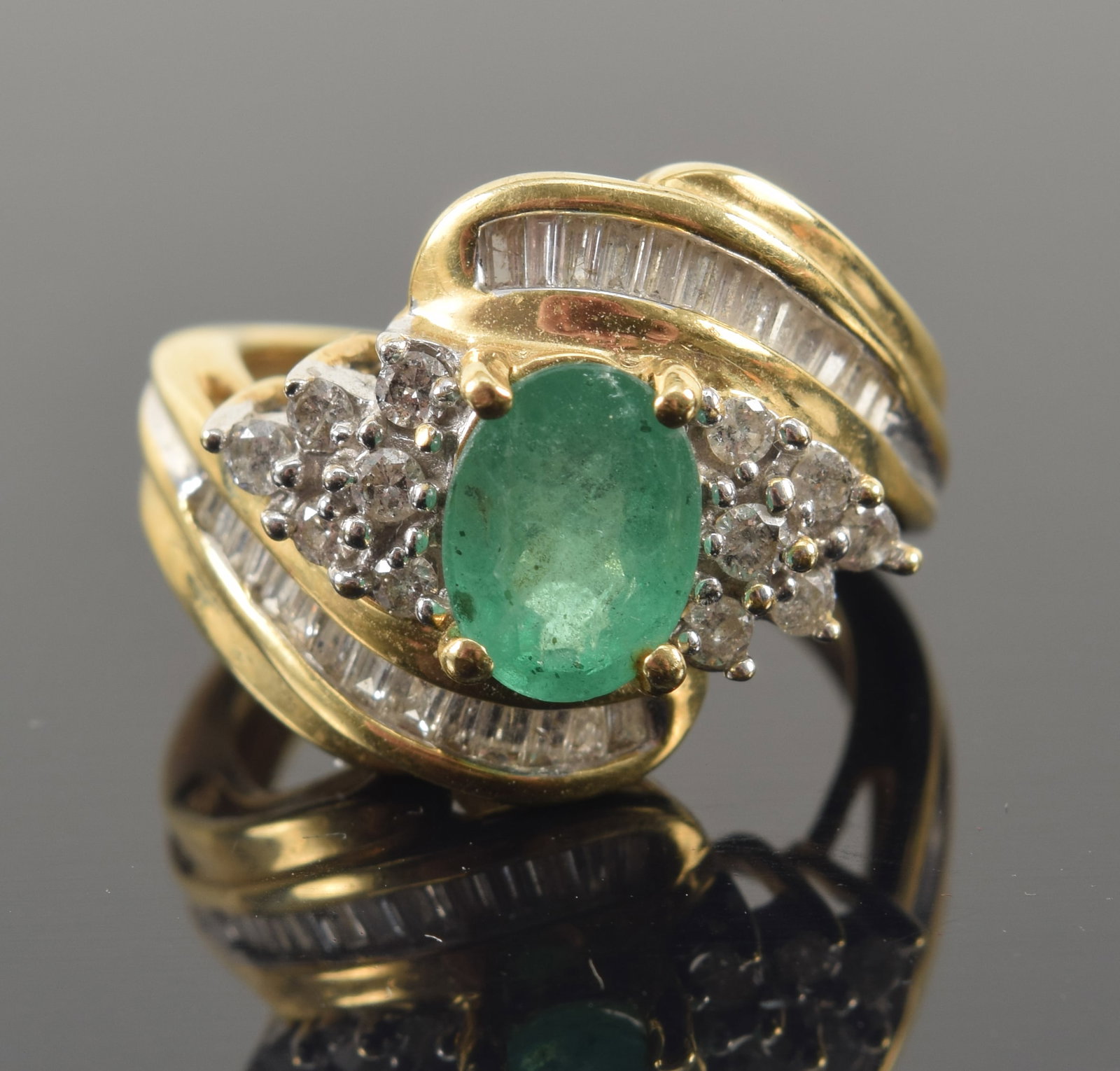 14K gold emerald and diamond bypass ring. Oval cut emerald measures approx. 7.8 x 5.9mm. Small: 14K gold emerald and diamond bypass ring. Oval cut emerald measures approx. 7.8 x 5.9mm. Small diamond clusters set in white gold and 2 channels of baguette cut diamonds. Marked 14K. Ring size 5. 5.3 