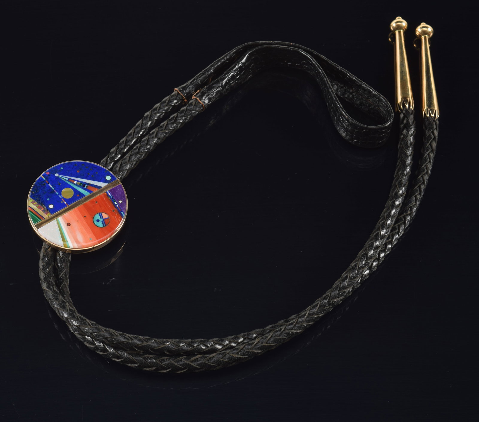 Jesse Monongya. Hopi-Dine. Gold inlaid bolo tie. Disc with mosaic stone inlay depicting space scenes: Jesse Monongya. Hopi-Dine. Gold inlaid bolo tie. Disc with mosaic stone inlay depicting space scenes from the artist's cosmos series. Braided leather tassels capped in gold. Stamped on the reverse. Th