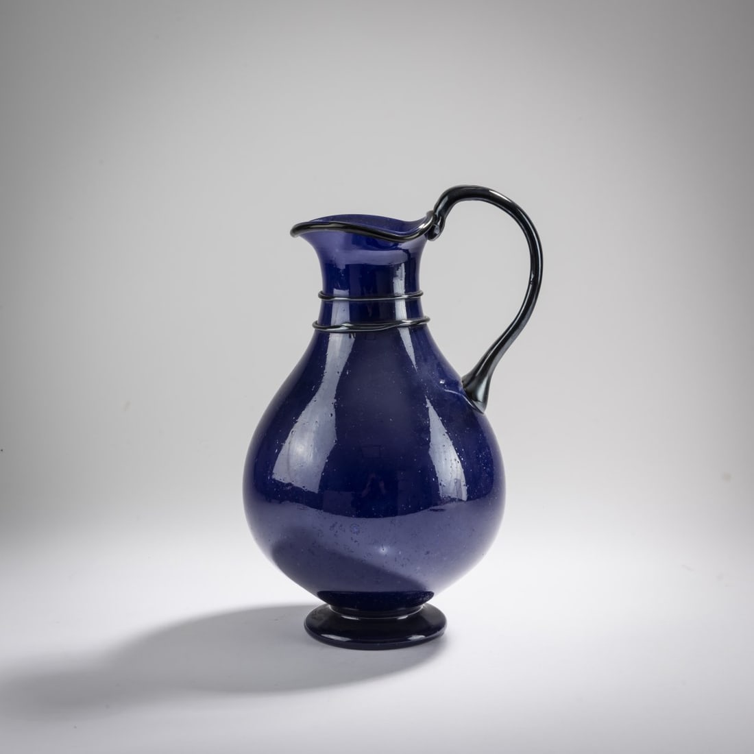 Napoleone Martinuzzi (attributed), 'Pulegoso' decanter, c. 1934: <p>H. 28 cm. Execution: Zecchin-Martinuzzi (attributed). Heavily bubbly blue glass, two applied threads, lip and handle deep purple; iridescent. Marked: C.V.M. (acid stamp).</p>