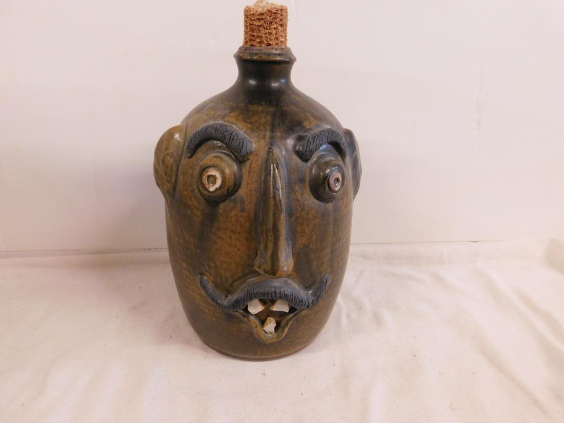 Sid Luck Signed Face Jug: Sid Luck Signed Face Jug, 1993, streaky brown glazed pottery, white glazed teeth and eyes, applied charcoal glazed eyebrows and mustache, corn cob stopper, 10 1/2" h. CONDITION: Very fine.