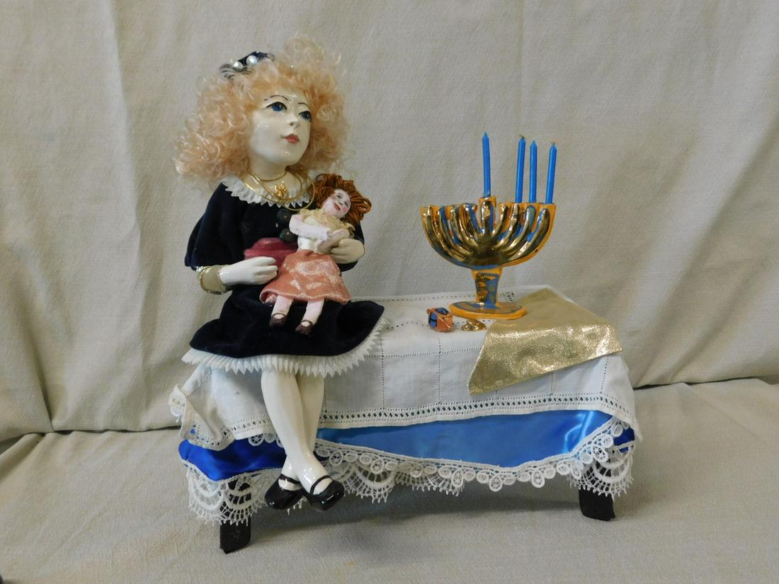 Barbara Kozero Judaica Sculpture: Barbara Kozero Judaica Sculpture, Pa artist, ceramic and cloth, depicts a ceramic girl, cloth dress holding a doll and sitting upon a cloth covered ceramic table that holds a menorah, Dreidel and coin