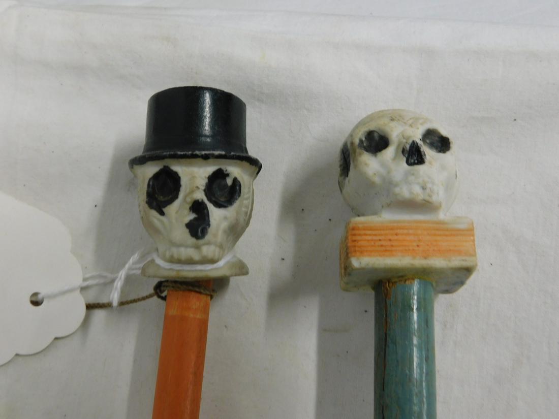 2 Skull Carnival Canes: Two Skull Carnival Canes, painted bisque skull head handles, one rests on a book the other wears a to9p hat and is marked "Japan", painted dowel shafts with metal ferrules, circa 1940s-50s, 34"l. [96]