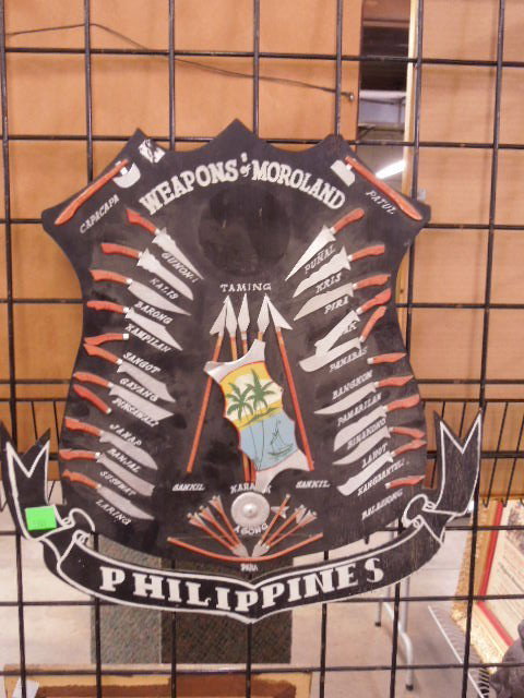 "Weapons of Moroland-Philippines" wooden Display: "Weapons of Moroland-Philippines" wooden display. Depicts various types of bladed hand weapons. Hand made on plywood backing. Measures: 22 1/2" x 21 1/2". CONDITION: Fair, missing pieces.