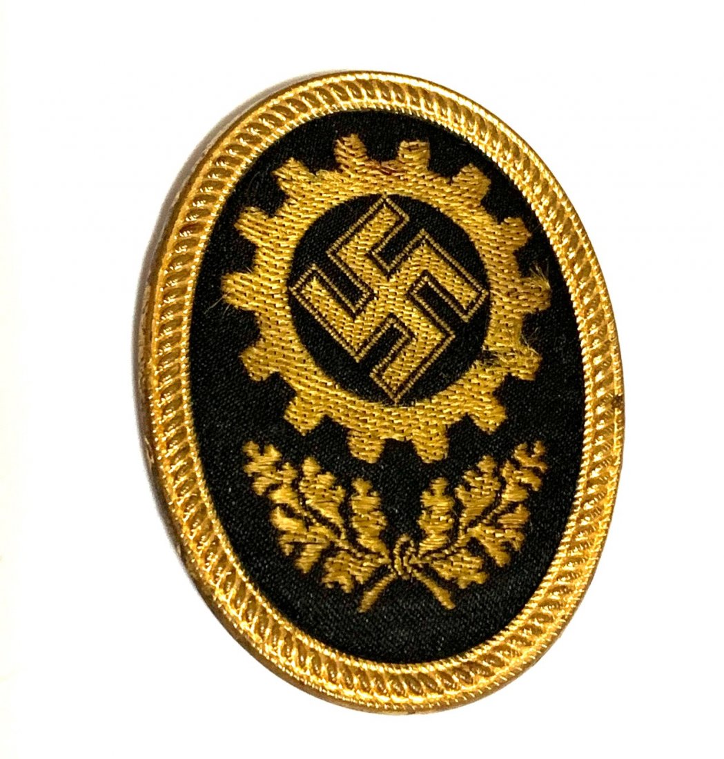DAF VISOR CAP BADGE: DAF Visor Cap Badge-the oval badge is made out of black and yellow cotton cloth with a die stamped gold gilded tombak exterior and die stamped steel based and silver washed backing. Obvrse dipicts a D