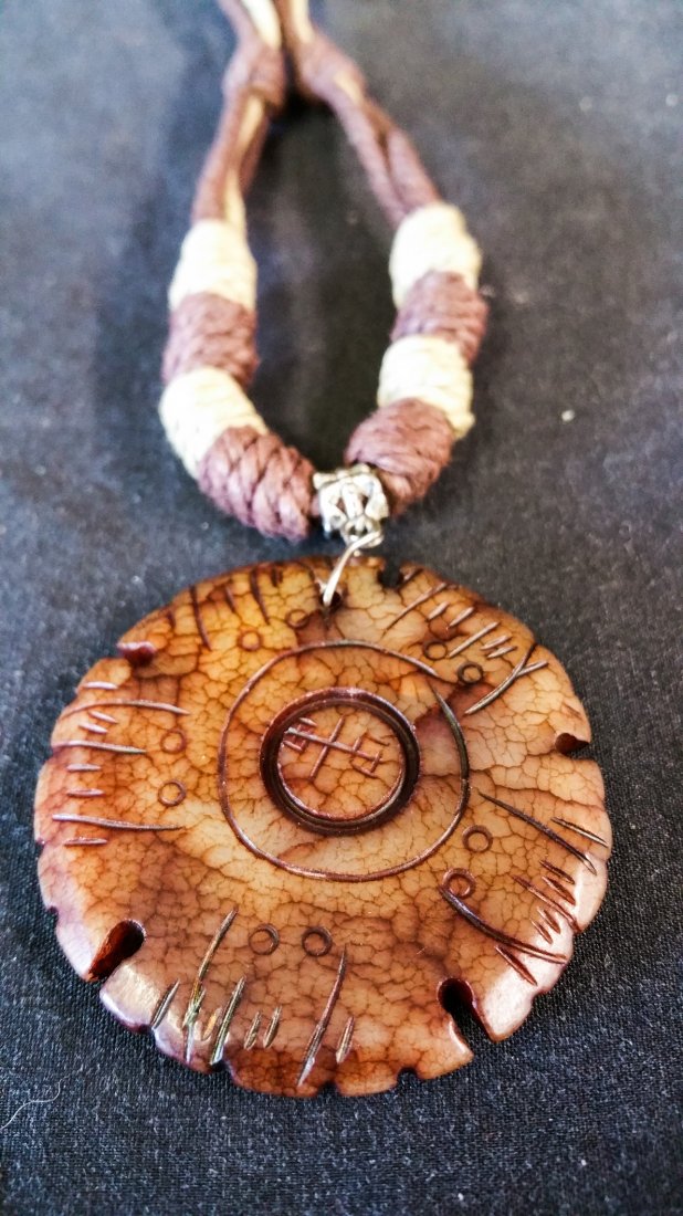 CARVED BROWN VEINED STONE DISC MEDALLION: North American carved brown veined partially translucent stone disc medallion, etched with an alternating line pattern and central glyph figure. 2 inch diameter disc on 24 in natural fiber knotted cor