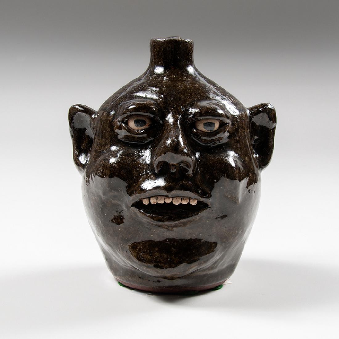 Lanier Meaders Face Jug: American, 20th century. A face jug by Lanier Meaders (1917-1998) in a dark alkaline olive glaze with stone teeth and painted ceramic eyes, signed on underside; ht. 8.25, wd. 7.25, dp. 6.5 in.