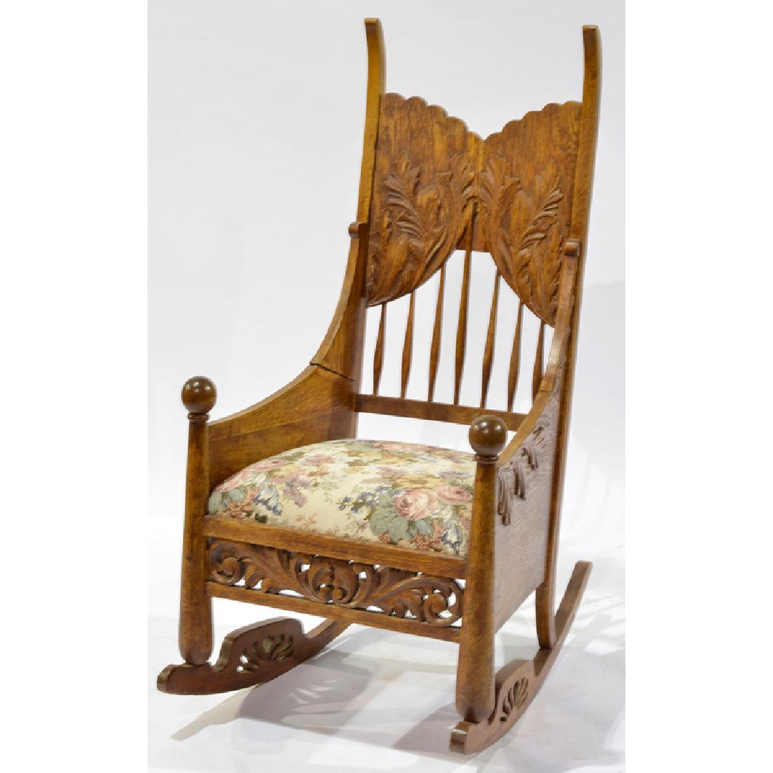 Folk Carved Rocking Chair: 20th century. A rocking chair in oak with foliate carving and ball handles, having an embroidered seat; oah. 49.25 in.