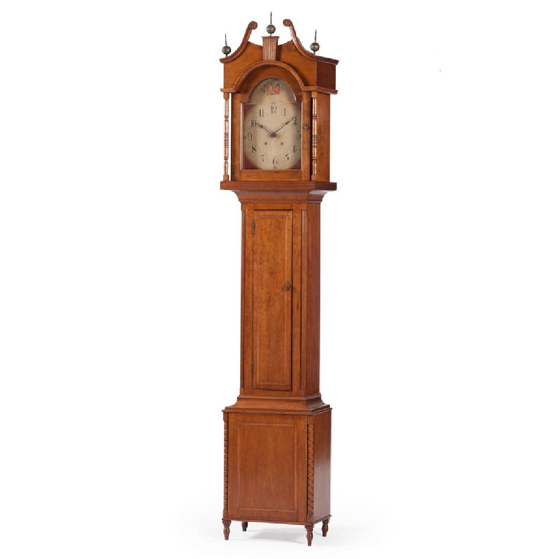 American Tall Case Clock in Cherry: American, 19th century. A tall case clock in figured cherry, having a hand-painted, floral decorated face with wooden works, the fluted and dovetailed bonnet with a broken arch pediment ending in chip