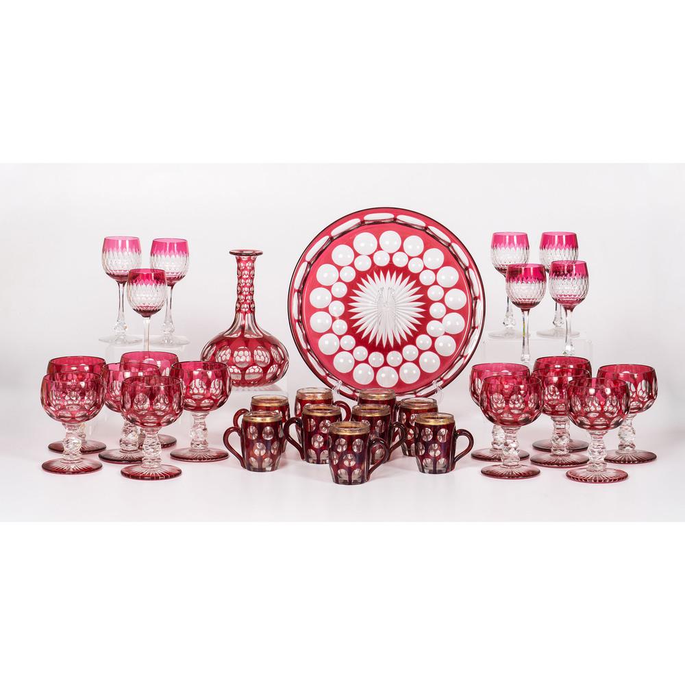 An Assortment of Ruby Cut to Clear Tableware: Likely Continental, 20th Century includes twelve water goblets, nine barrel-shaped cups, a platter and decanter in the Blarney pattern, and seven glasses. Height of decanter 8 3/4 inches. Diameter of