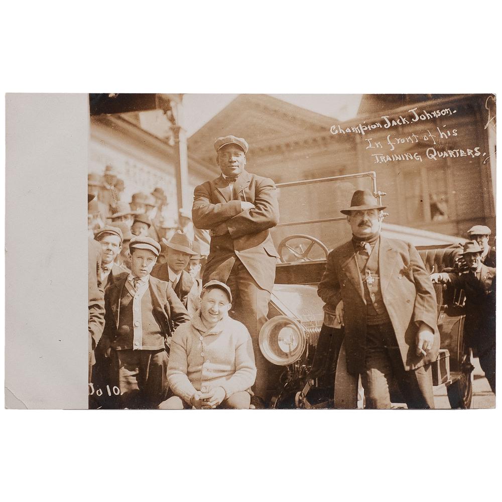 Champion Jack Johnson in Front of his Training: Real photo postcard showing a confident Jack Johnson with his retinue. 5.5 x 3.5 in. Captioned in negative: "Champion Jack Johnson. In front of his Training Quarters" alongside an apparent date "Ju 1