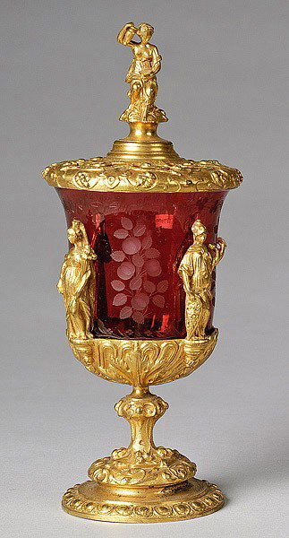 GILT BRONZE & CRANBERRY GLASS CHALICE : GILT BRONZE & CRANBERRY GLASS CHALICE Continental. An etched cranberry center vase in a ormolu mount with classical figures and finial. 9" high.