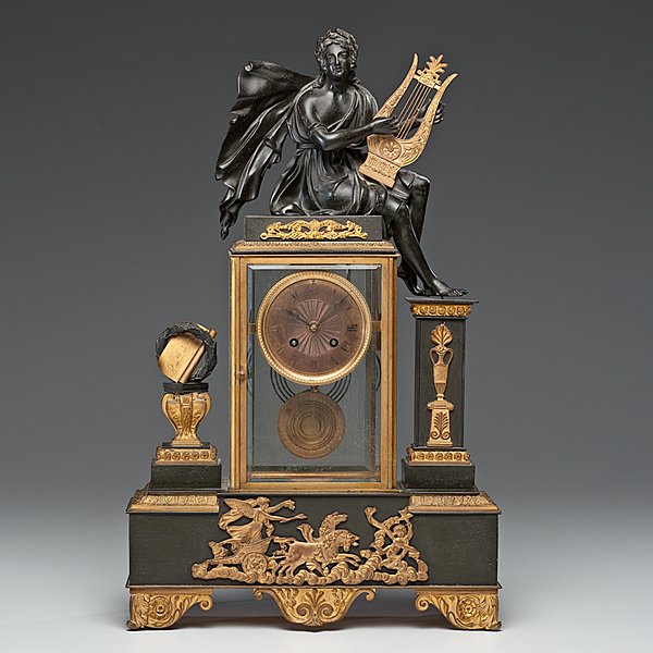 French Mantel Clock : French Mantel Clock French. A bronze and ormolu mantel clock, with a glass case with seated allegorical robed figure holding a lyre, the case flanked by an urn and column, the base with a chario