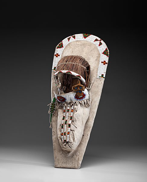 Ute Beaded Hide Cradleboard Collected by John S.: sinew-sewn on hide and beaded using colors of red, green, orange, faceted rose, red, blue, and white; miniature moccasins and gauntlets hang from basketry visor; lizard umbilical charm hangs from sid